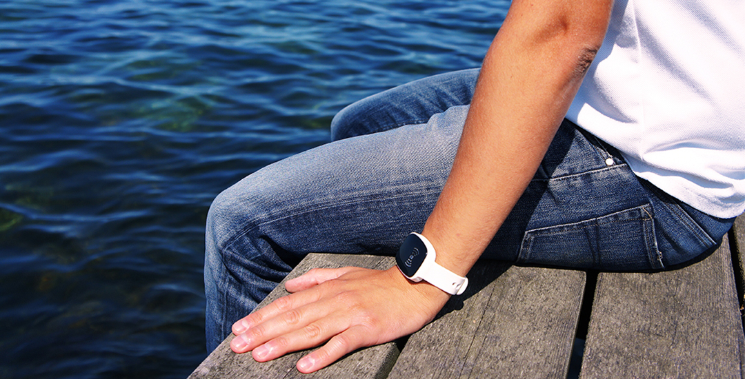 Person sitting on a wooden dock with one hand resting beside them, wearing a white smart wristband with a visible screen. The wristband stands out against denim jeans and the deep blue water, suggesting reliability in outdoor, waterside settings.